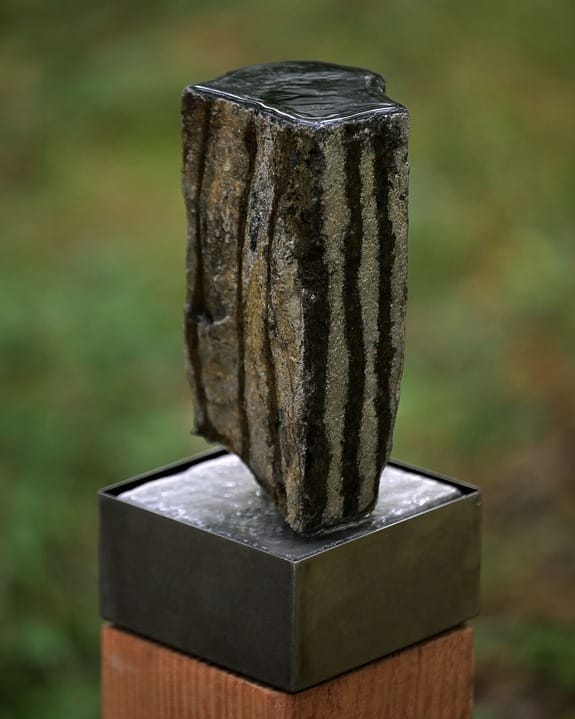 Vertical stone sculpture with layered textures on a metal pedestal, set against a blurred green background.