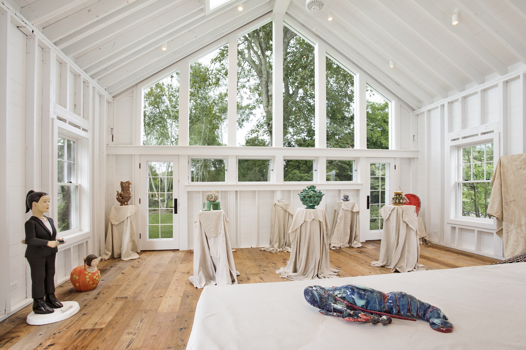 Art gallery with sculptures on pedestals covered in cloth, large windows, wooden floor, and a view of trees outside.