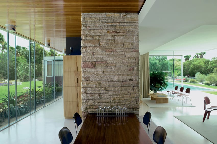 Modern living area with stone wall, wooden table, and large windows overlooking a garden and pool.