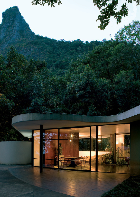 Modern glass-walled building with scenic mountain backdrop, surrounded by lush greenery and trees, under a clear sky.