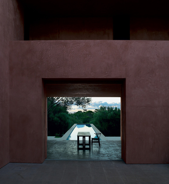 A minimalist outdoor patio with a table and chair overlooking a serene pool and lush greenery under a cloudy sky.