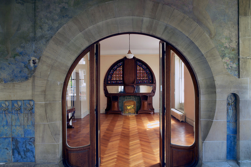 Elegant room with ornate wooden furniture, arched stone entryway, and decorative stained glass window in a historic building.