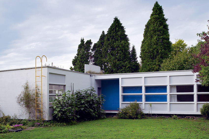Modern white house with large windows surrounded by lush greenery and tall trees on a cloudy day.