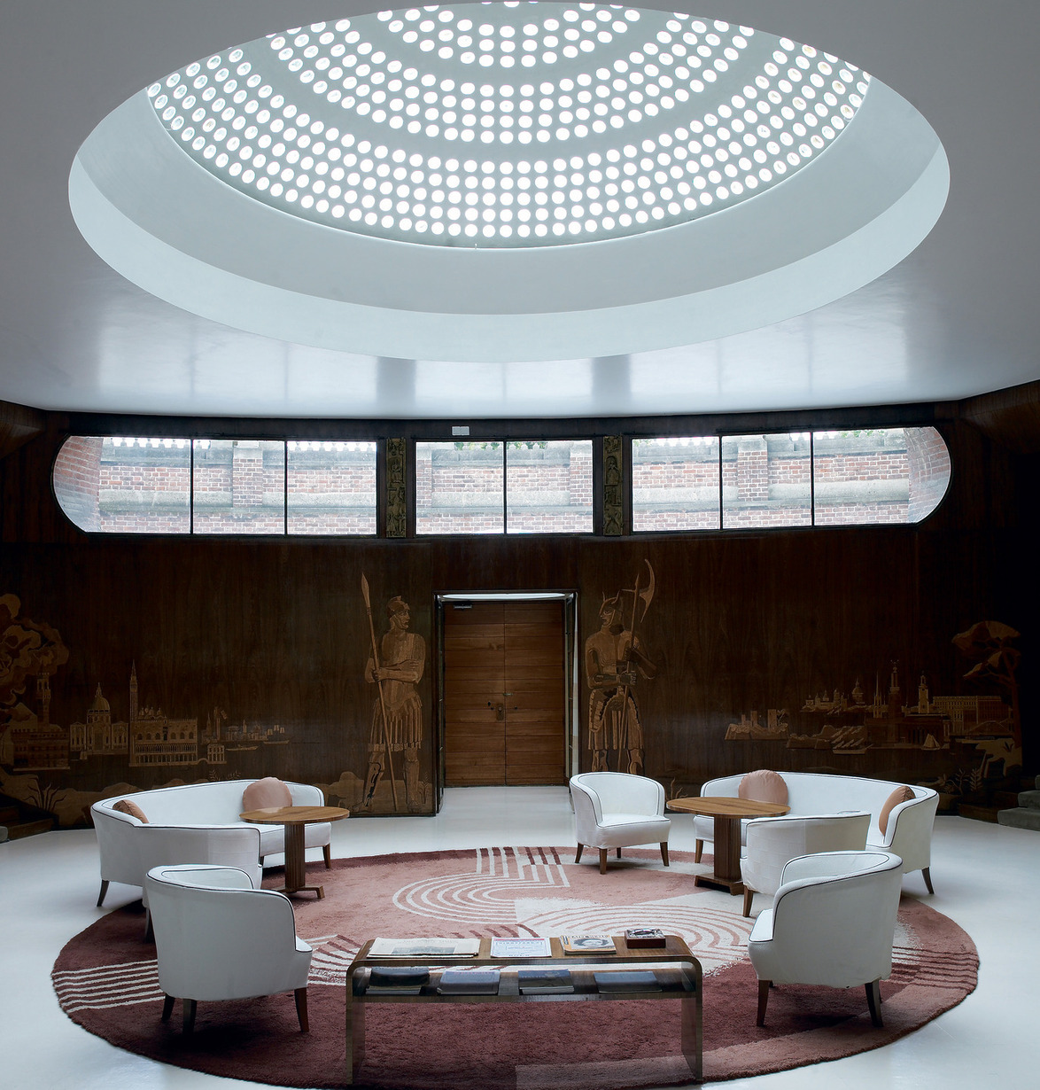 Modern circular room with round skylight, white chairs on a patterned rug, and wooden wall art in the background.