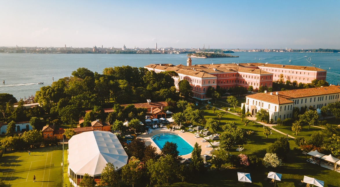 Aerial view of a resort with a pool and lush gardens by the sea, with a distant cityscape under a clear blue sky.