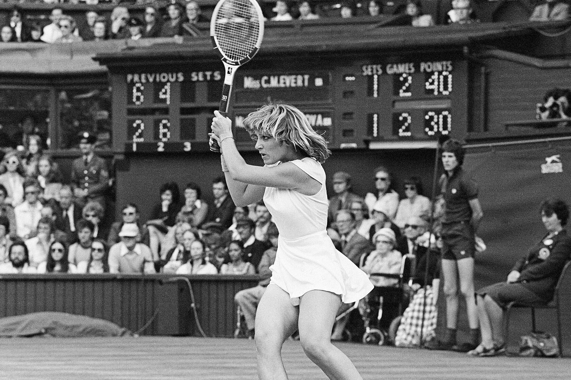 Tennis player mid-swing during a match with spectators and a scoreboard in the background at a tournament.