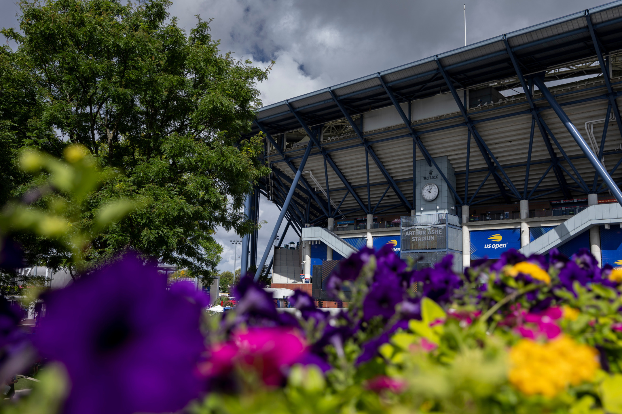 Arthur Ashe Stadium with vibrant flowers in the foreground under a cloudy sky.