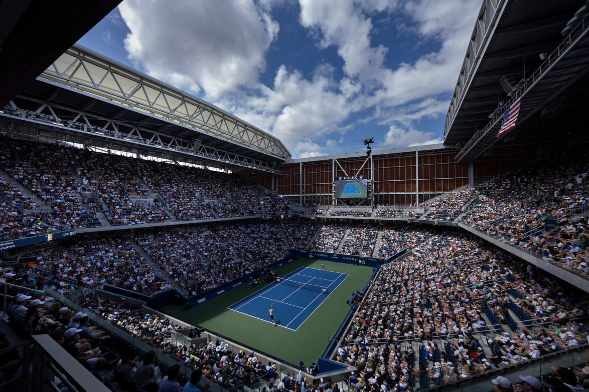 Crowded tennis stadium with a match in progress under a partly cloudy sky.