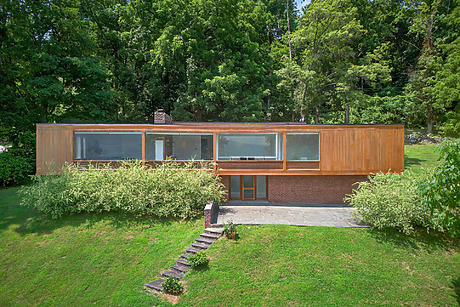 Modern wooden house with large windows surrounded by greenery and trees on a sloped grassy hillside.
