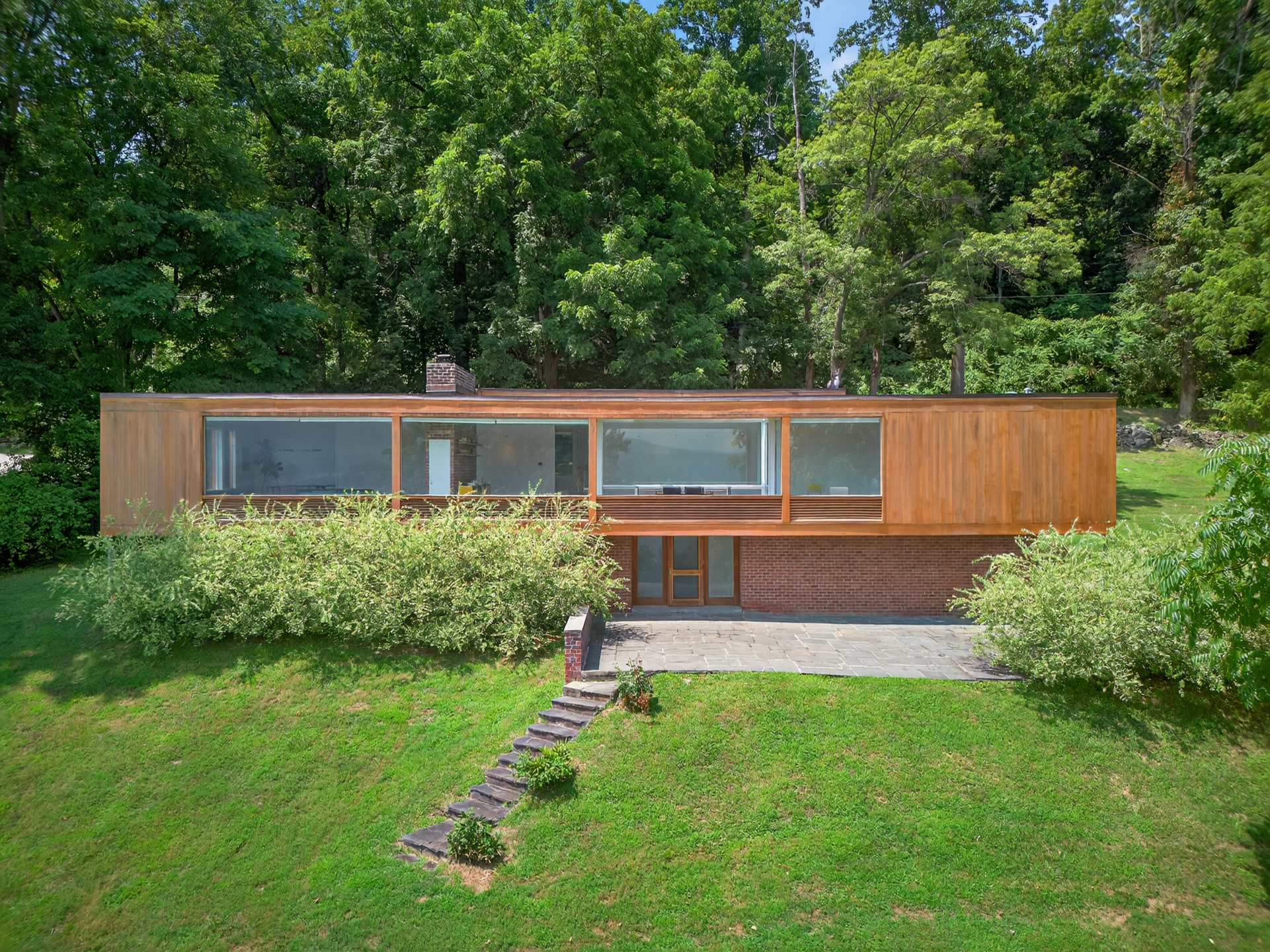 Modern wooden house with large windows surrounded by greenery and trees on a sloped grassy hillside.