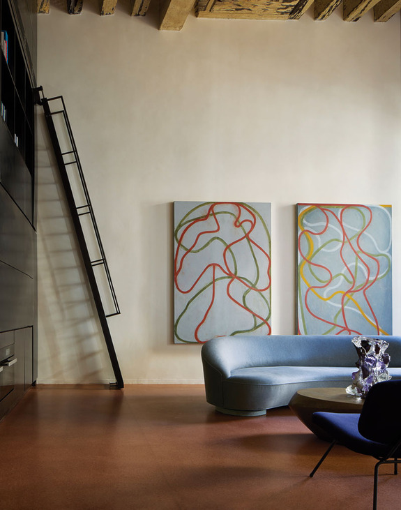 Modern living room with abstract art, blue curved sofa, black shelving, and a ladder against a beige wall.