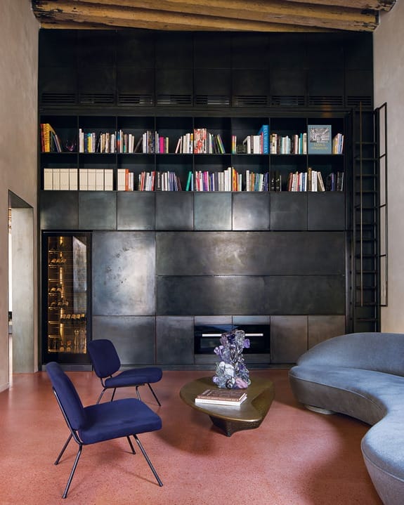 Modern living room with dark bookcase, blue chairs, curved gray sofa, wooden coffee table, and red terrazzo floor.