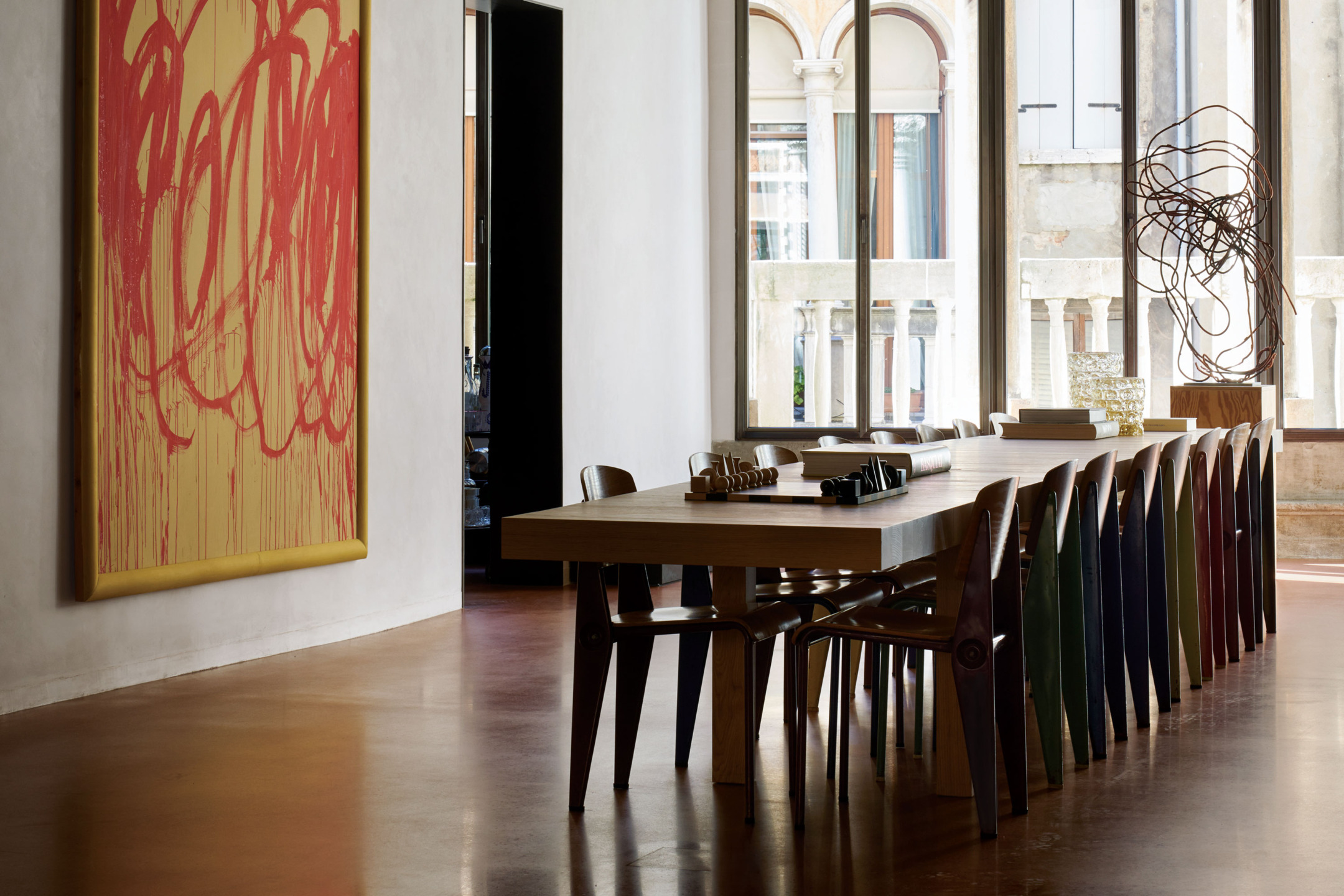 Modern dining room with a long wooden table, colorful chairs, abstract art on wall, and large windows letting in natural light.