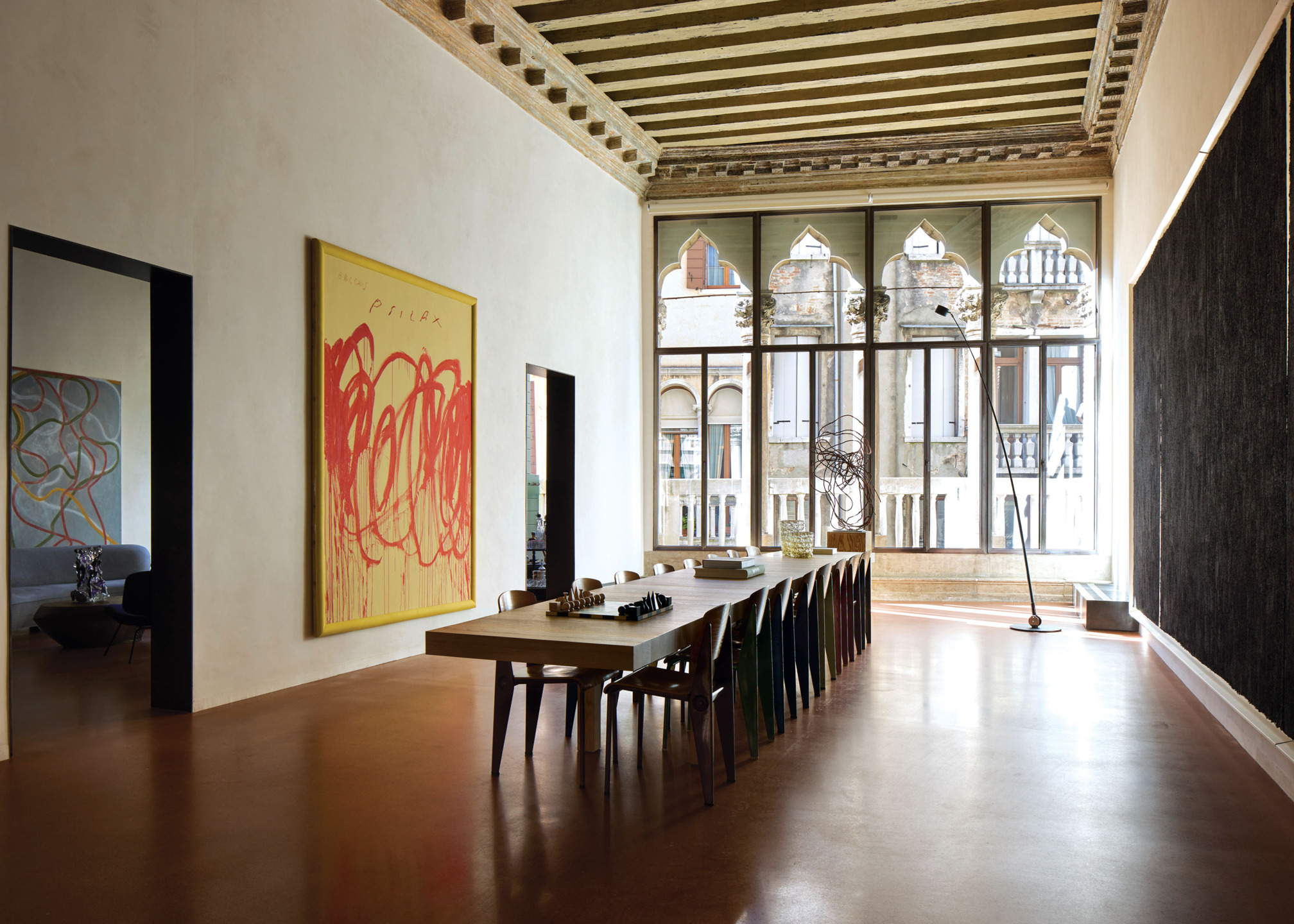 Spacious dining room with a long table, modern art on walls, large windows, and wooden ceiling beams.