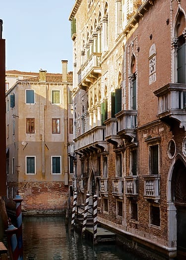 Historic Venetian building along a narrow canal lit by afternoon sunlight, showcasing ornate balconies and architecture.