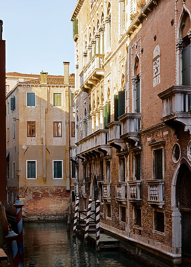 Historic Venetian building along a narrow canal lit by afternoon sunlight, showcasing ornate balconies and architecture.