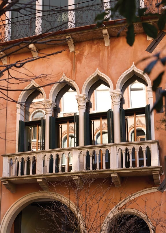 Ornate arched windows of an Italian building with open shutters and a small balcony surrounded by tree branches.