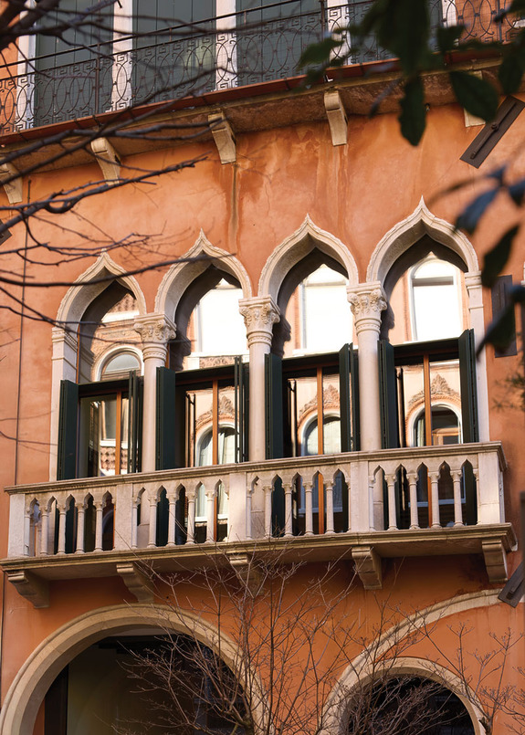 Ornate arched windows of an Italian building with open shutters and a small balcony surrounded by tree branches.