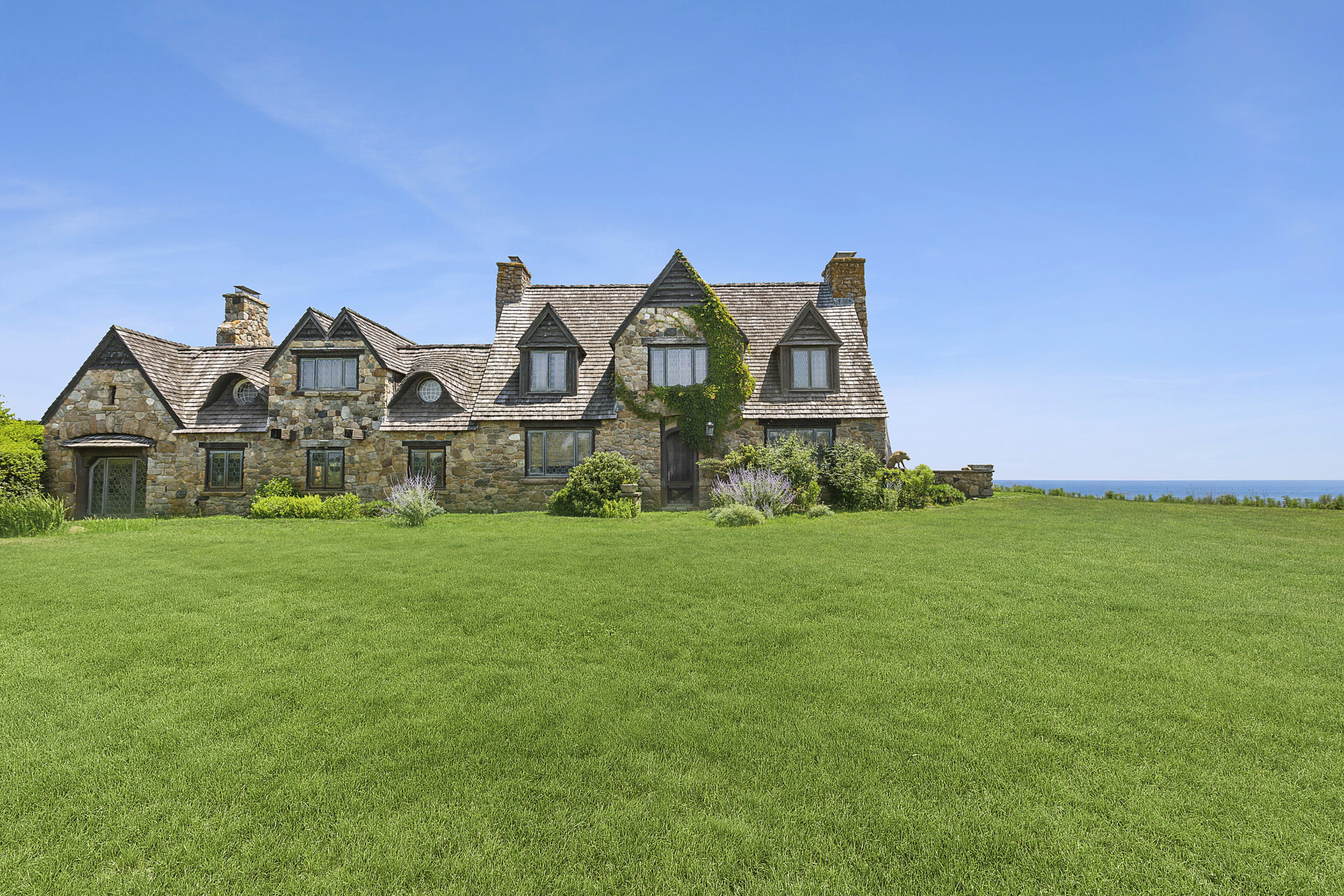 Stone cottage with a steep roof and greenery on a large grassy lawn under a clear blue sky.