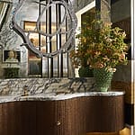Luxurious bathroom with curved wood vanity, large marble backsplash, abstract mirror, and vibrant floral arrangement.