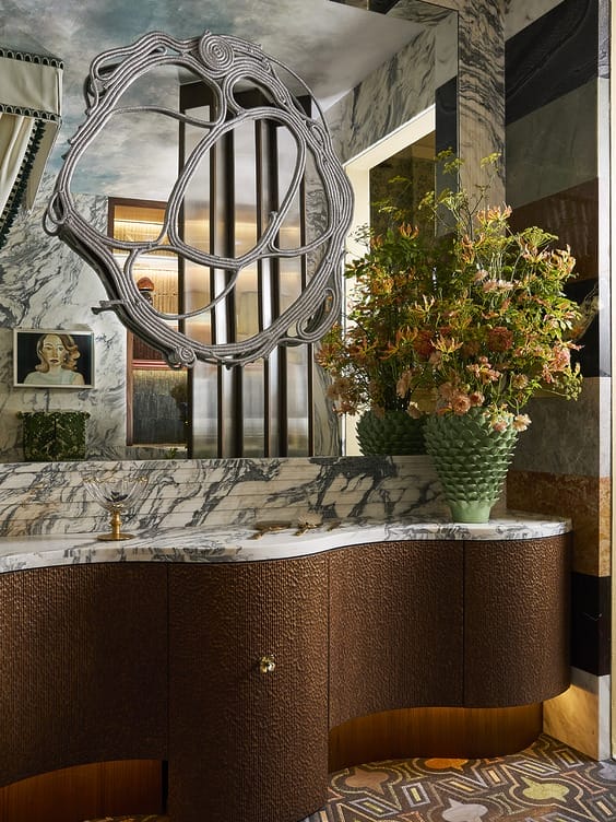 Luxurious bathroom with curved wood vanity, large marble backsplash, abstract mirror, and vibrant floral arrangement.