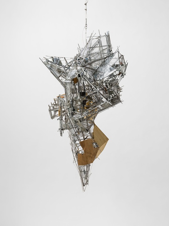 Abstract metal sculpture suspended against a plain background, composed of jagged, reflective metal pieces and wooden elements.