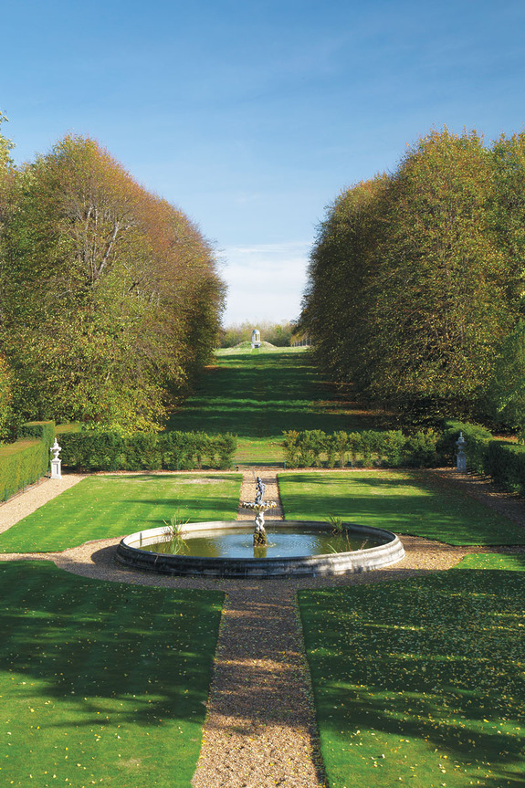 Formal garden with a circular fountain, lined with neatly trimmed hedges and trees under a clear blue sky.