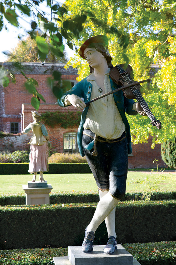 statue of a man playing violin in garden with vibrant green trees and historic brick building in background