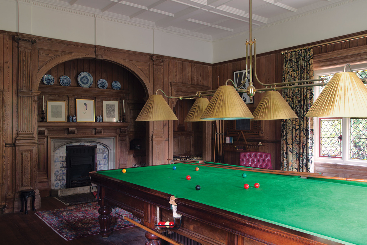 Luxurious billiard room with a green pool table, ornate wooden walls, large fireplace, and classic hanging lights.