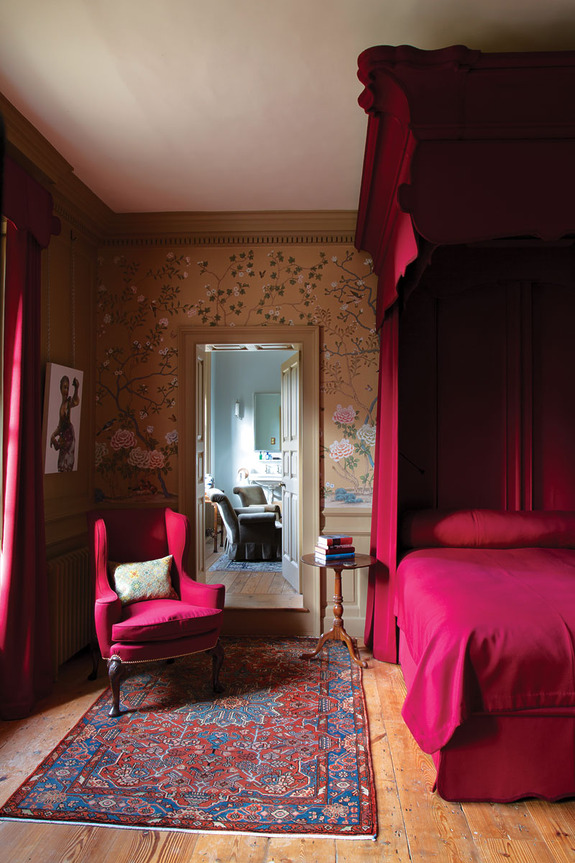 Elegant bedroom with red accents, canopy bed, decorative wallpaper, armchair, colorful rug, and view into an adjoining room.