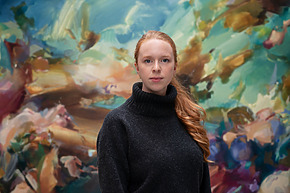 Person with long red hair wearing a dark sweater stands in front of a colorful abstract mural.