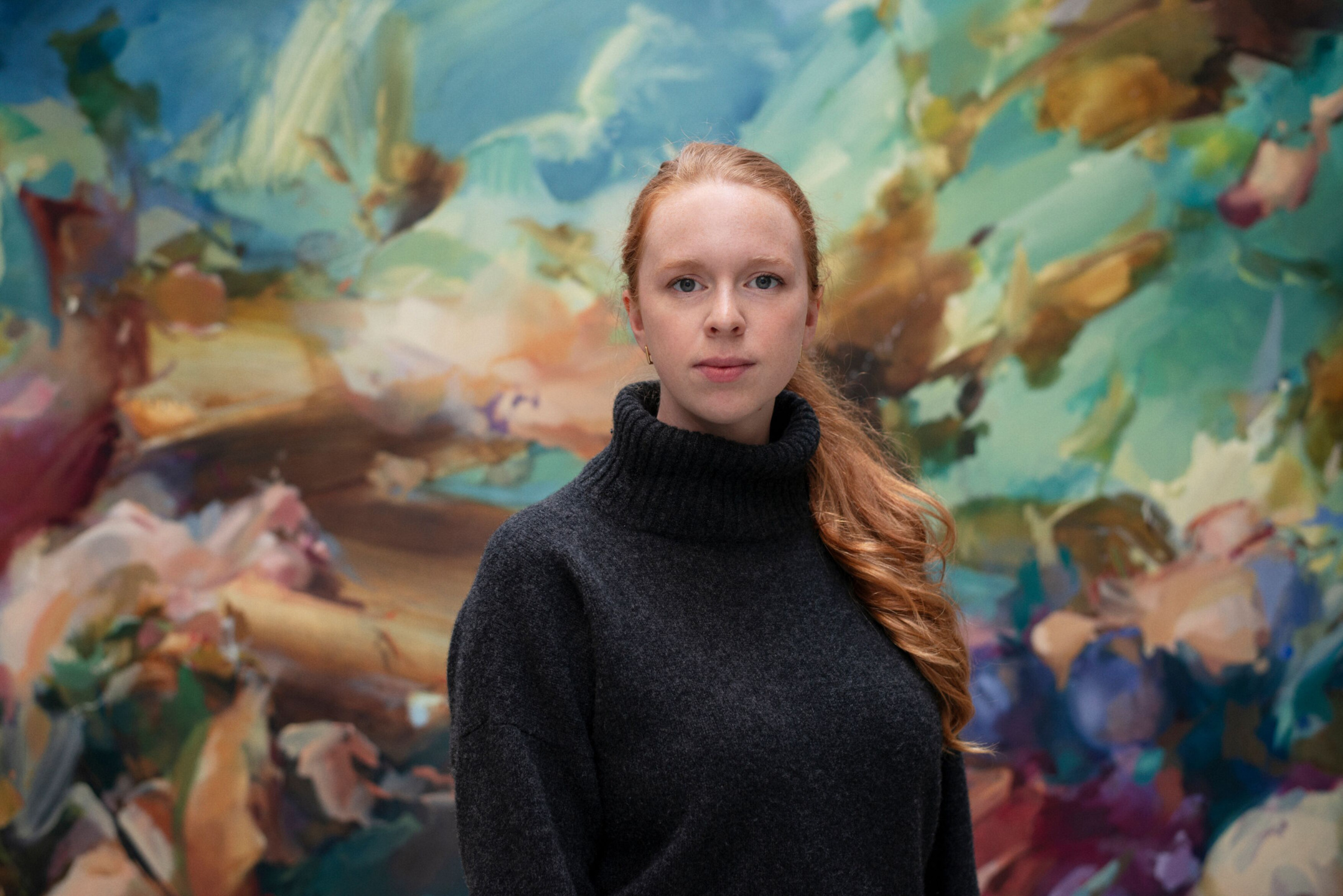 Person with long red hair wearing a dark sweater stands in front of a colorful abstract mural.
