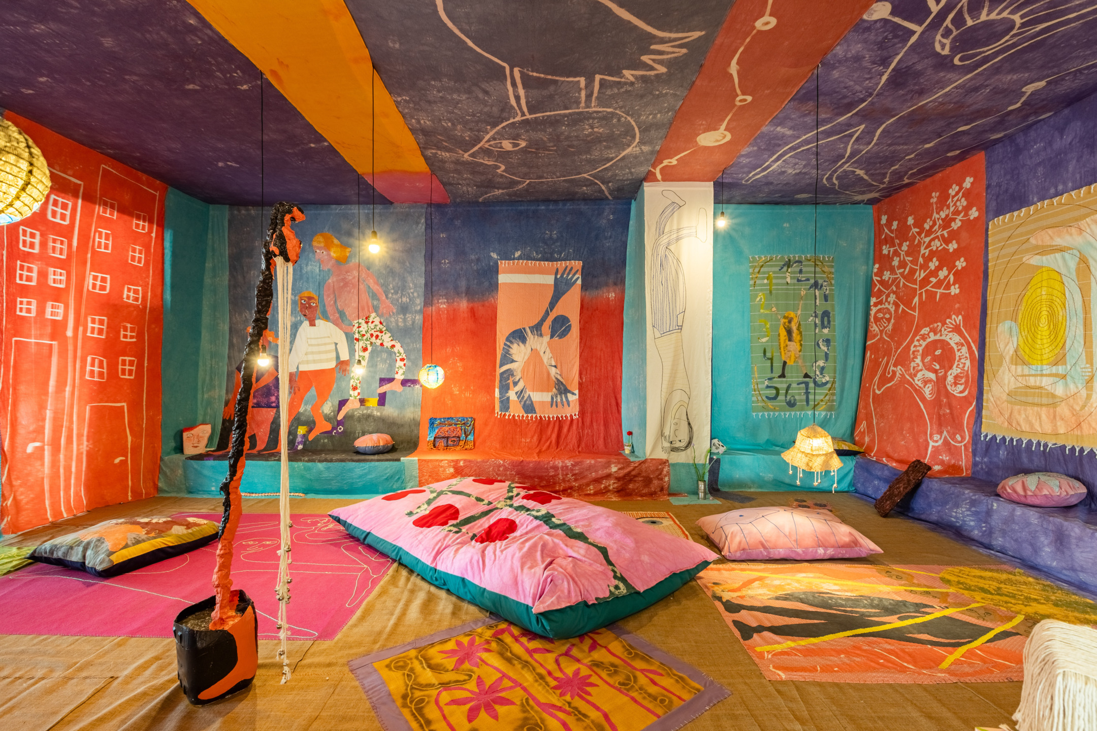 Colorful room with artistic murals on walls and ceiling, decorated with cushions and hanging lights.