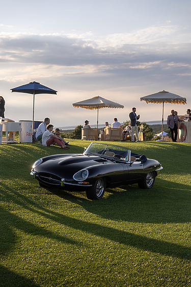 Vintage black convertible car on a grassy field with people relaxing under umbrellas in the background at sunset.