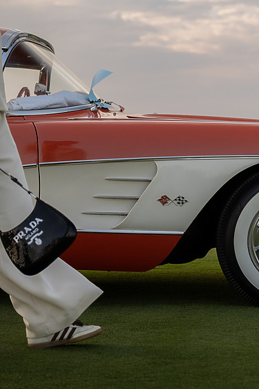 Person walking by a vintage red and white car with a Prada bag, wearing white pants and striped shoes on grassy field.