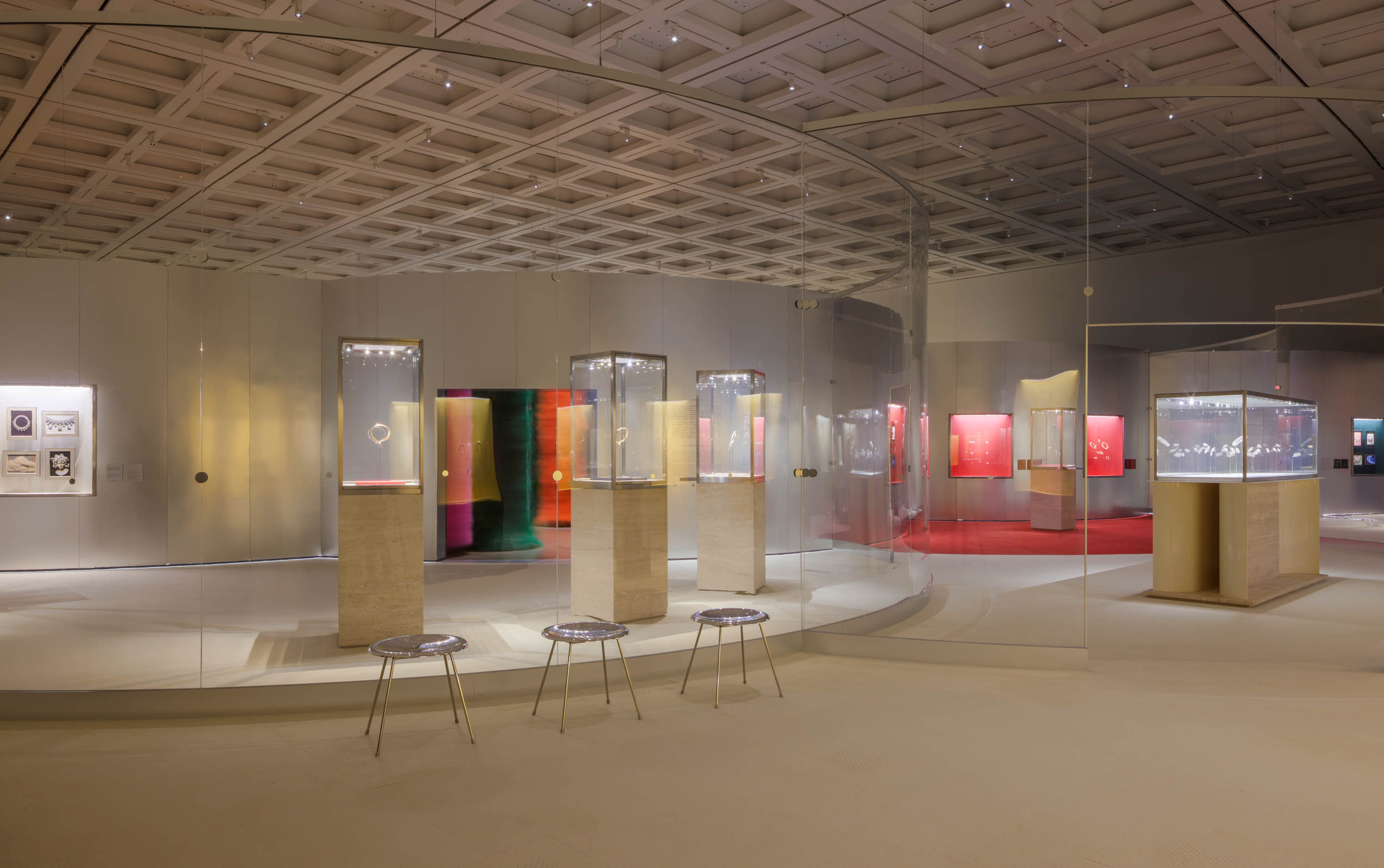 Modern art gallery interior with illuminated display cases, vibrant paintings, stools, and a geometric ceiling.