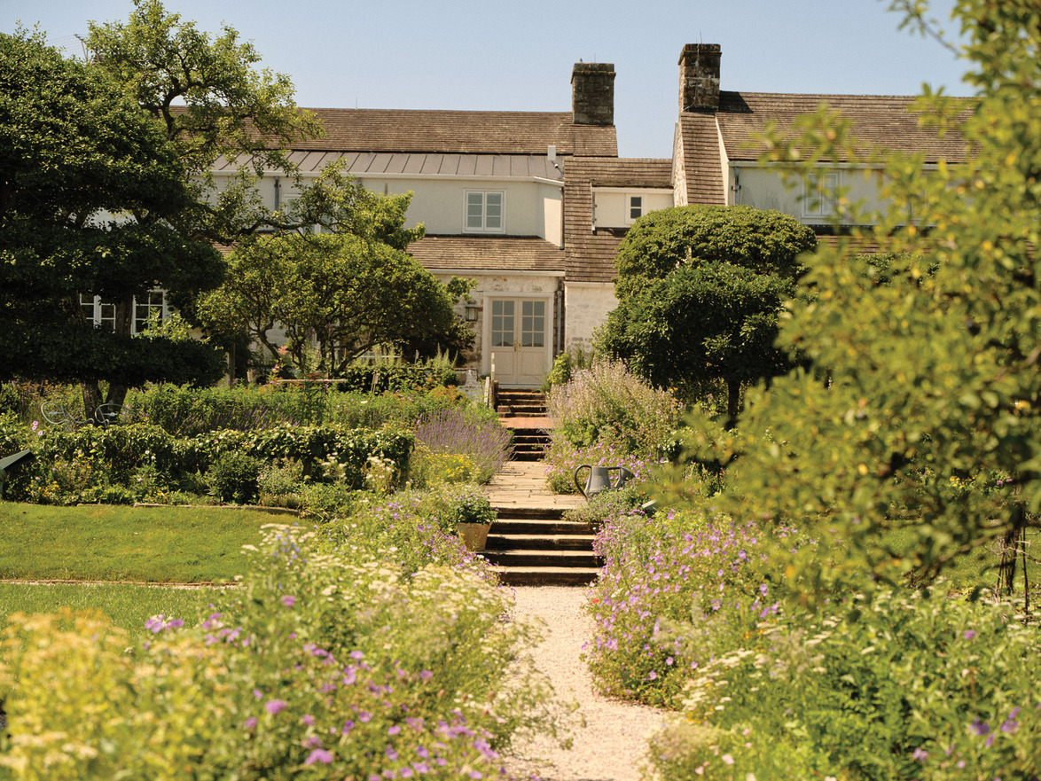 Pathway leading through a lush garden towards a large, elegant house with multiple chimneys in the background