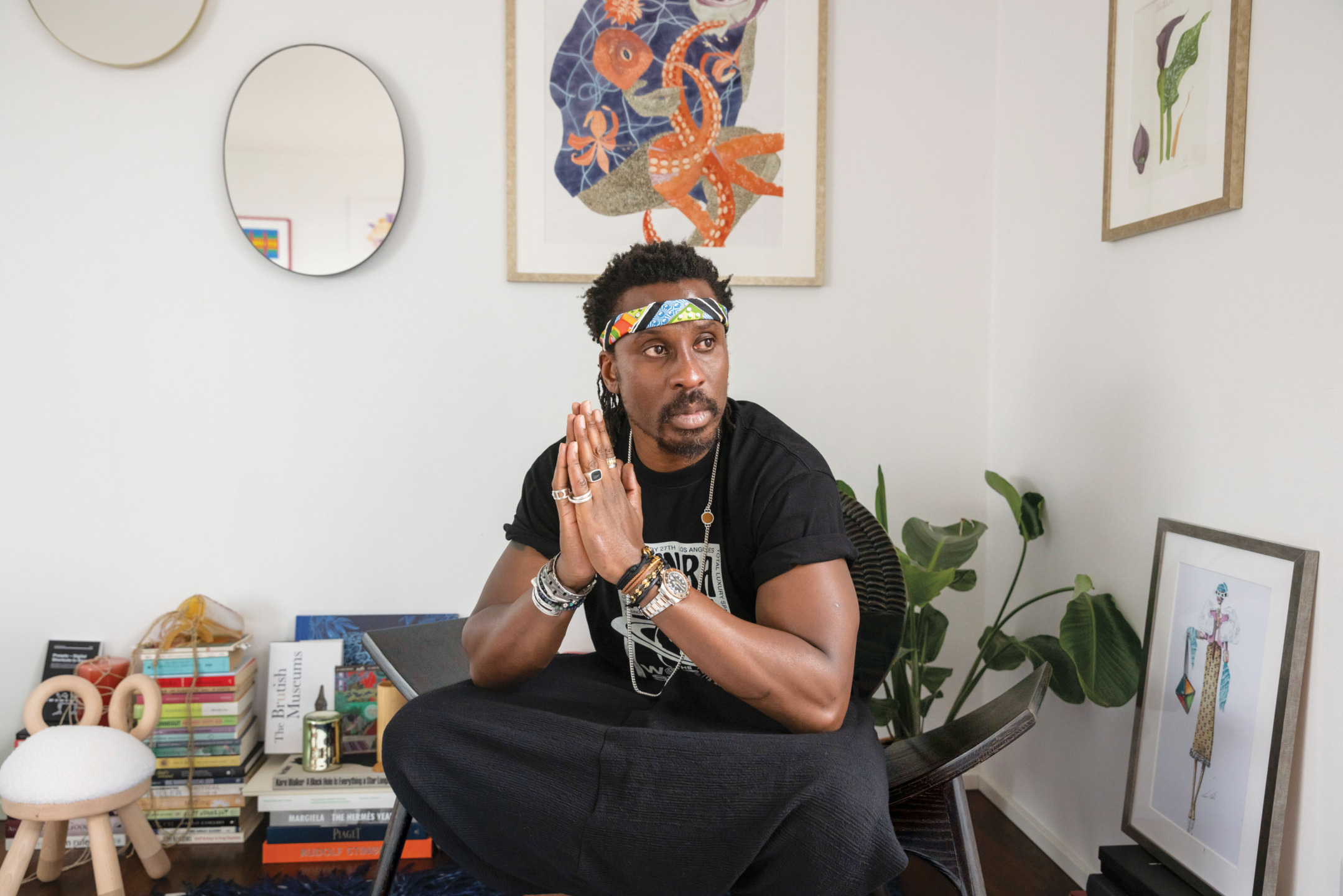 Man sitting in a thoughtfully decorated room with art, books, and plants, wearing a colorful headband and rings.