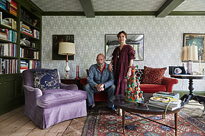 Two people sitting and standing in a cozy living room with colorful furniture, bookshelves, and decorative items.