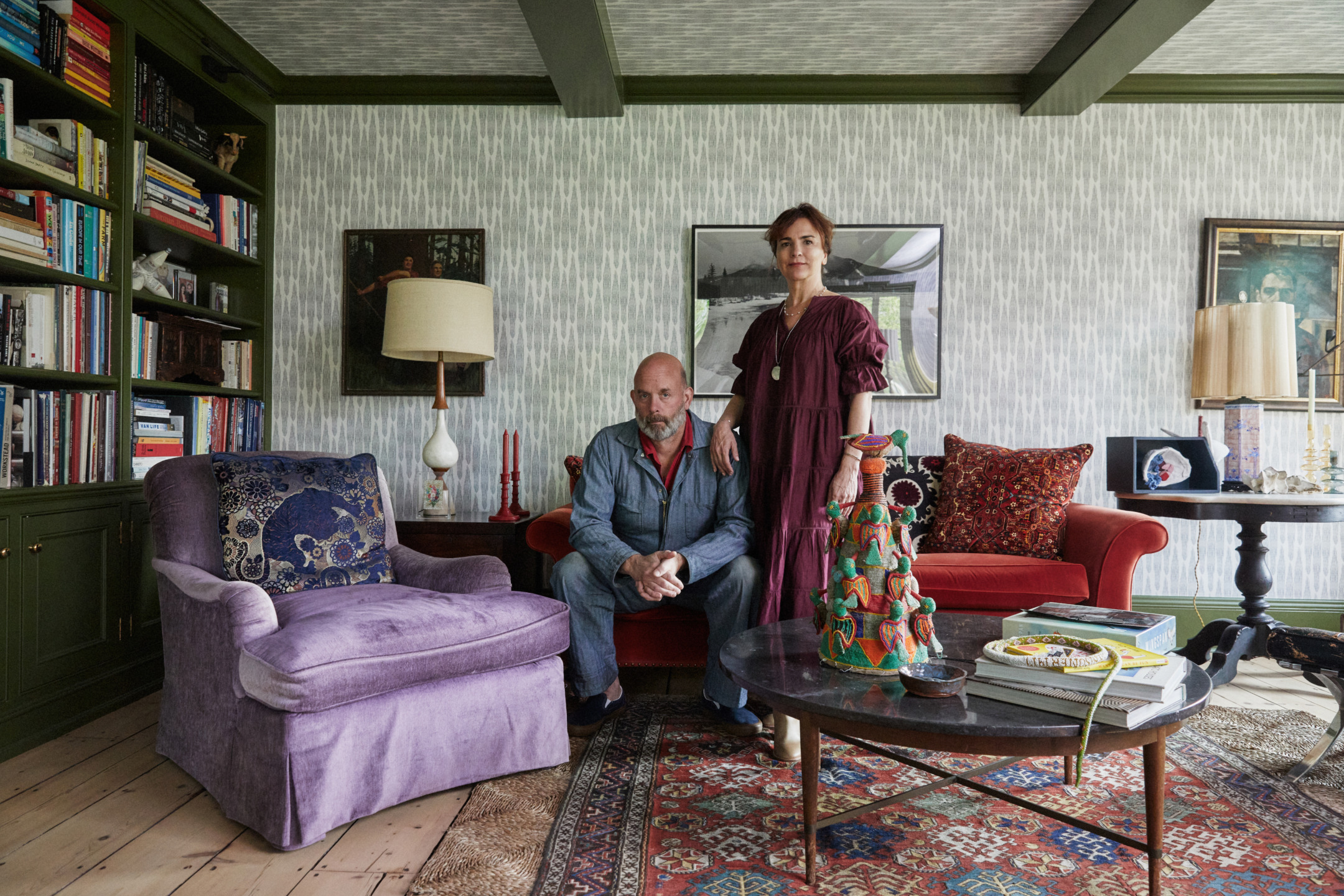 Two people sitting and standing in a cozy living room with colorful furniture, bookshelves, and decorative items.