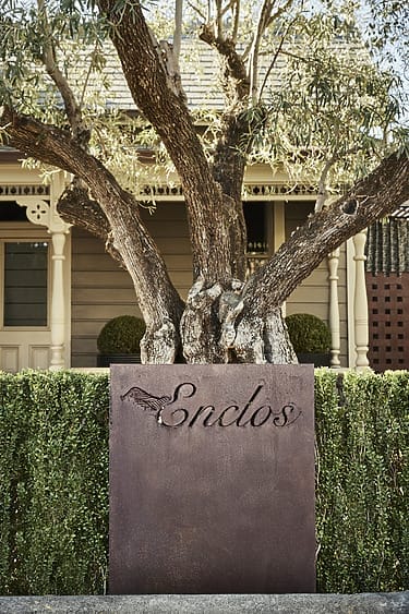 Rustic sign with the word "Enclos" in front of a tree and a traditional house.
