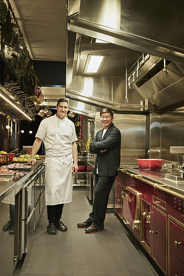 Two men standing in a modern kitchen with stainless steel appliances and a red oven, one wearing a chef's uniform.