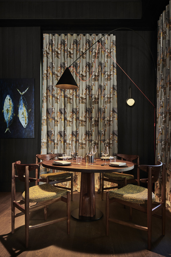 Cozy dining area with a round table, four wooden chairs, and elegant curtains in a dimly lit room with wall art.