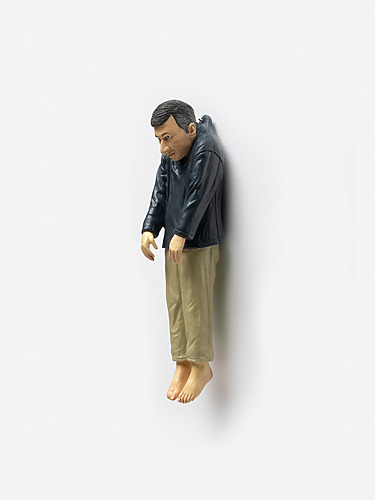 Small sculpture of a man with a dark jacket and beige pants, leaning against a white wall, appearing thoughtful.