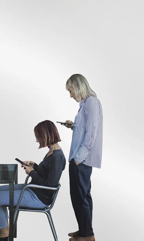 Two people using smartphones, one sitting at a table and the other standing, both focused on their devices.