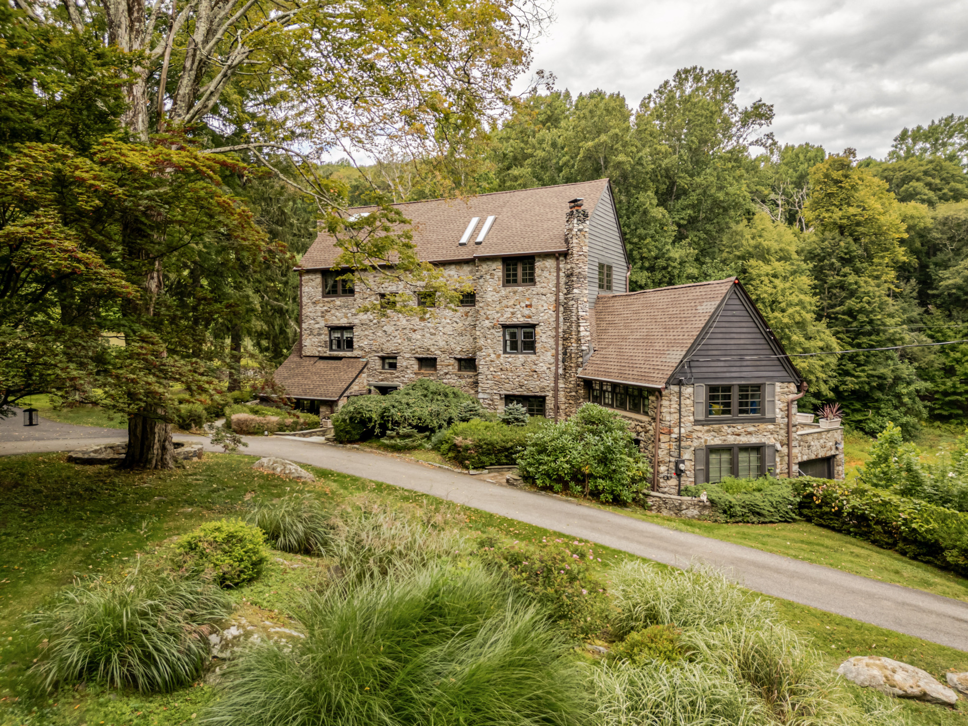 Stone house surrounded by lush greenery and trees, with a sloped driveway leading to the entrance, under a cloudy sky.