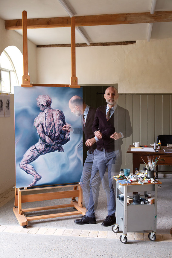 Artist standing beside a large canvas on an easel, painting supplies on a cart next to him, in a well-lit studio.