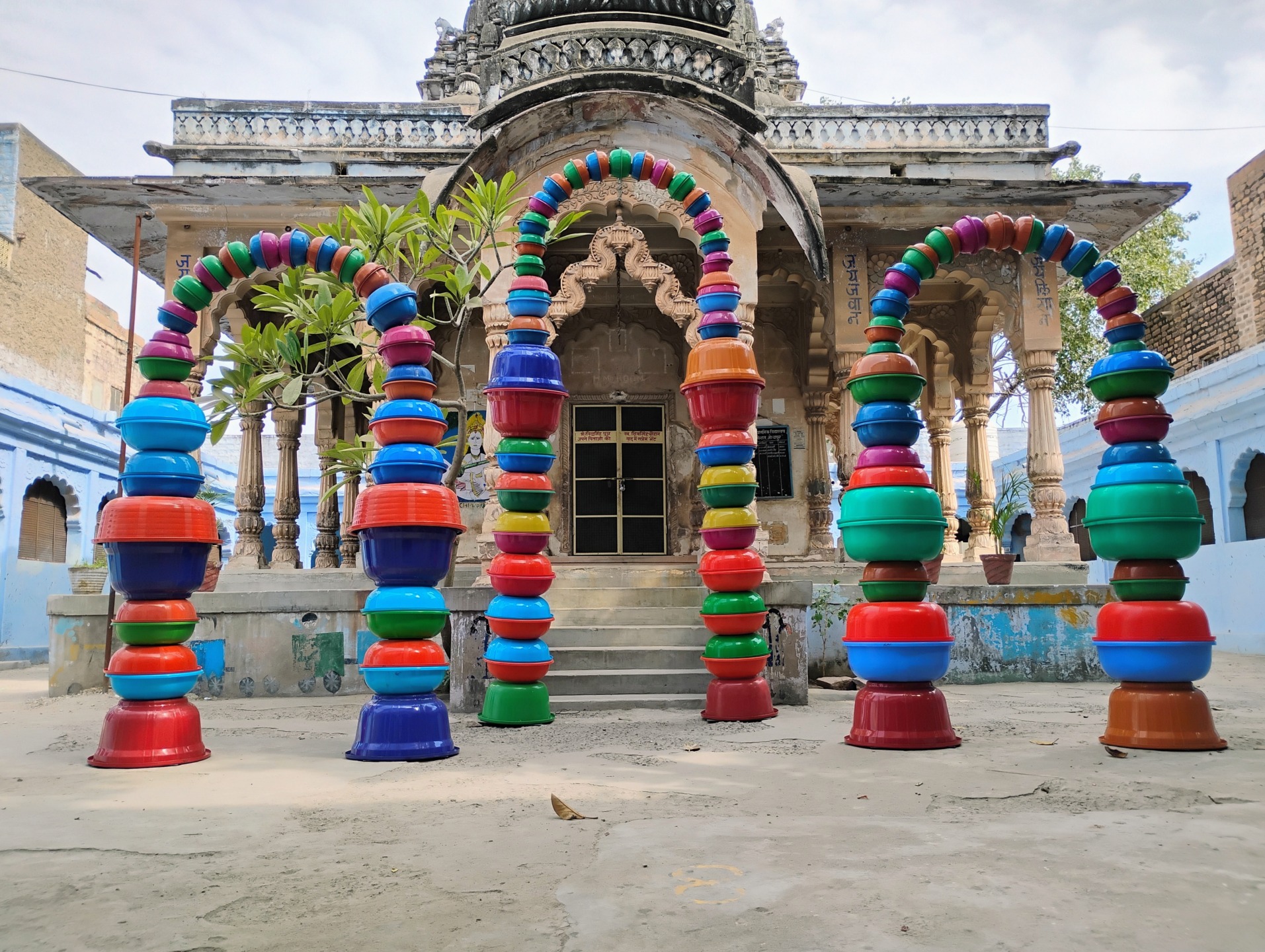 Colorful arches made of stacked bowls in front of a historic building with intricate architecture and surrounding trees.