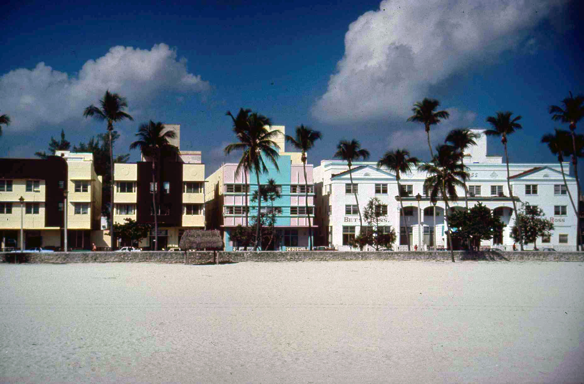 Colorful Art Deco buildings with palm trees lining a sandy beach under a partly cloudy sky.