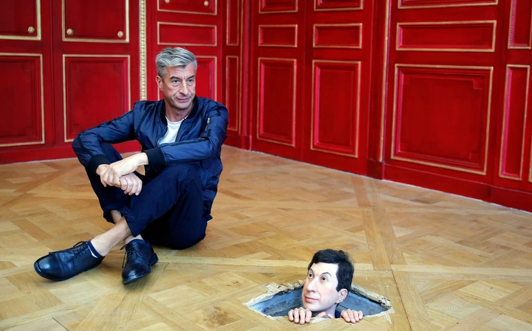 Man sitting on a wooden floor next to a realistic sculpture of a man's head emerging through a hole in the floor.